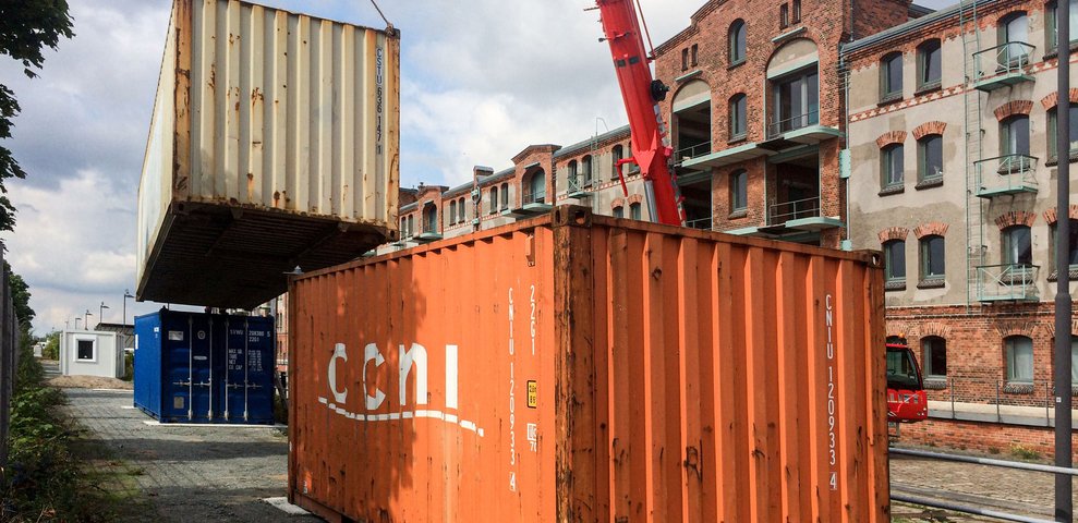 Kran hebt Schiffscontainer neben orangefarbenem Container am Speicher XI, Backsteingebäude dahinter.