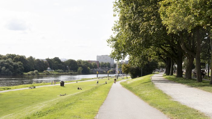 Bremer Osterdeich, Flusspfad mit üppigen grünen Wiesen und Bäumen, Menschen spazieren und radeln an einem sonnigen Tag