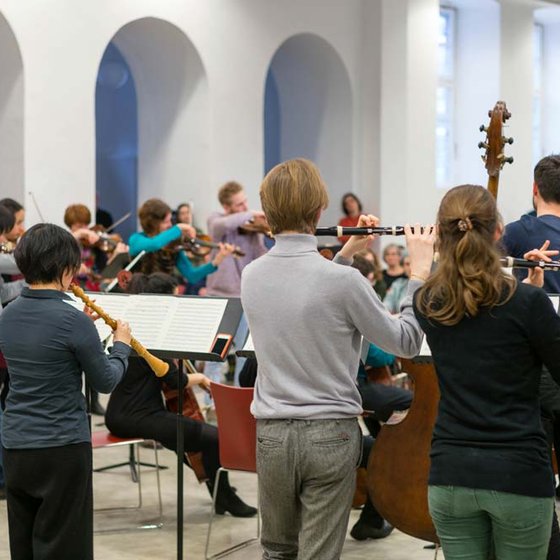 Musiker:innen spielen in einem geräumigen Raum, spielen verschiedene Instrumente, mit Blick auf den Rücken eines Flötisten und andere Streicher, Publikum im Hintergrund